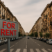 Cartello For Rent in primo piano su strada urbana con palazzi residenziali – simbolo del calo della domanda di affitti nelle città italiane nel 2025
