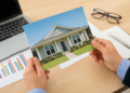 Mani che tengono una foto di una casa moderna, con grafici e laptop su scrivania, simbolo di analisi delle tendenze mercato immobiliare 2025
