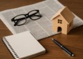 Modello di casa in legno, giornale, taccuino e occhiali su scrivania: concetto di notizie immobiliari